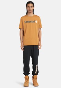 T-shirt marrone Timberland con logo, abbinata a pantaloni sportivi neri con branding stampato e stivali beige con lacci e suole a contrasto.