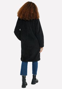 Cappotto nero peloso dal design semplice, tessuto testurizzato e silhouette dritta, abbinato a denim blu corto e stivali neri.