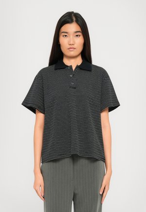 Young woman with long black hair wearing a black and white striped polo shirt and gray pinstriped pants, standing against a plain background.