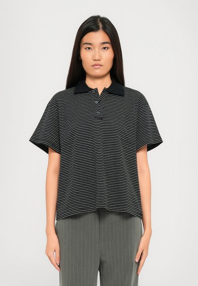 Young woman with long black hair wearing a black and white striped polo shirt and gray pinstriped pants, standing against a plain background.
