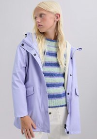 Persona joven con cabello largo y rubio que lleva una chaqueta lila claro sobre un suéter a rayas azul y verde y pantalones blancos, sobre un fondo liso.