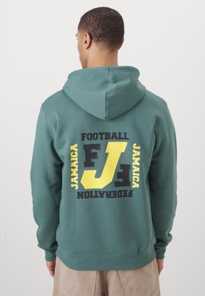 Mand iført grøn hættetrøje med gul og sort "JAMAICA FOOTBALL FEDERATION" tekst og stort stiliseret "FJF" logo på ryggen.
