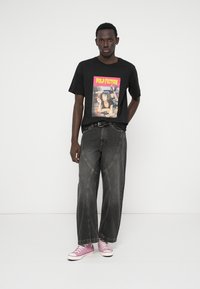 Νέος άνδρας που φοράει μαύρο t-shirt με γραφικά από το "Pulp Fiction", ξεθωριασμένα μαύρα τζιν, ροζ ψηλές αθλητικές γόβες, που στέκεται μπροστά σε άσπρο φόντο.