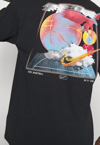 T-shirt noir arborant un motif graphique coloré représentant un ballon de basket, des nuages et un oiseau, avec le texte "NIKE BASKETBALL NO FLY ZONE".