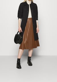 Cappotto di lana nero con bottoni, dolcevita crema, gonna plissettata marrone, stivaletti neri in pelle alla caviglia e una borsa nera texturizzata.