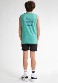 Turkis ærmeløs t-shirt med teksten "Key West Petrol Ind" i lilla, parret med sorte shorts og hvide strømper med sorte striber.