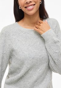 Γυναίκα με χαμόγελο που φορά ασημένες σκουλαρίκια και ένα ανοιχτό γκρι πουλόβερ με πλέξη, αγγίζοντας τον γιακά με το δεξί της χέρι.