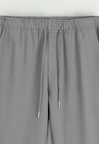 Pantalons à taille élastique gris clair avec cordons de serrage et poches latérales, montrant le détail avant supérieur sur un fond uni.