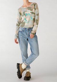 Maglione a maniche lunghe con motivo mimetico chiaro e una grafica di faccina sorridente turchese. Abbinato a jeans azzurri sdruciti e stivali marroni con lacci.