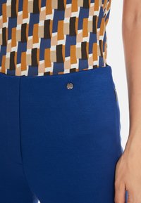 Blue fitted trousers with a smooth texture and a metal logo button on the waistband, paired with a patterned top showcasing blue, brown, and cream colors.