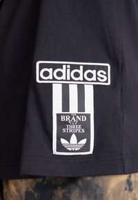 Μαύρη βαμβακερή μπλούζα t-shirt με λογότυπο "adidas" σε λευκό χρώμα, συνοδευόμενη από μια ορθογώνια ετικέτα που παρουσιάζει ένα σχέδιο τριών γραμμών και λεπτομέρειες του λογοτύπου.