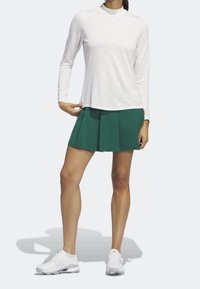 Bílý top s dlouhým rukávem s jemnou geometrickou texturou, kombinovaný se zelenou plisovanou sukní a bílými sportovními botami.