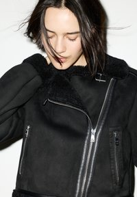 Giacca in suede nera con colletto in pelliccia, chiusura con zip e tasche laterali. Presenta una texture liscia e dettagli a zip neri a contrasto.