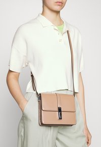 Křížová taška z opálené kůže s klopou, kontrastním černým kováním a hladkou strukturou. Kompaktní design s dlouhým nastavitelným popruhem.