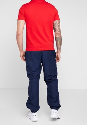 Chemise rouge à manches courtes avec un col rond, associée à un pantalon de jogging bleu marine, doté de poignets élastiques, et des chaussures de sport blanches.