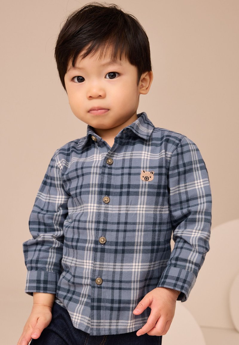 Chemise à carreaux bleue à boutons avec manches longues, présentant des motifs à carreaux bleu foncé et bleu clair et une petite broderie de cochon sur la poitrine.