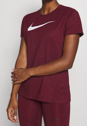 Person iført en burgunderfarvet Nike t-shirt og leggings, med den ene arm krydset over den anden mod en ensfarvet lys baggrund.