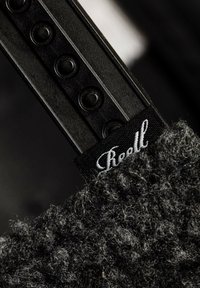 Schwarzer, strukturierten Stoff mit einem flauschigen Finish, angebracht an einem starren schwarzen Kunststoffriemen. Besitzt erhabene, kreisrunde Akzente und ein weißes Markenetikett.