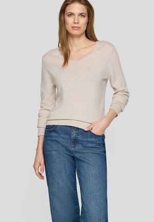 Woman standing with one hand in pocket, wearing a light beige V-neck sweater and blue jeans against a plain gray background.