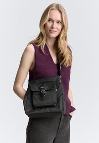 Zwarte stoffen tas met een voorvak, verstelbare schouderriem en metalen hardware. De tas wordt over de schouder gedragen voor een lichte achtergrond.