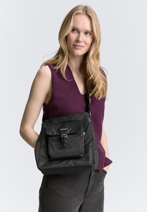 Schwarze Stofftasche mit einer Fronttasche, verstellbarem Riemen und Metallbeschlägen. Die Tasche wird über der Schulter vor einem hellen Hintergrund getragen.