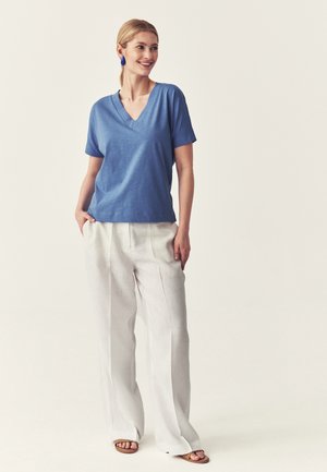 Kobieta uśmiechająca się, ubrana w niebieską bluzkę z krótkim rękawem i dekoltem w serek, białe spodnie z szerokimi nogawkami, niebieskie kolczyki oraz brązowe sandały, stoi z rękami w kieszeniach.