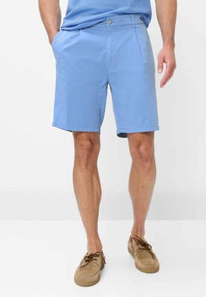 Mann trägt hellblaue, maßgeschneiderte Shorts und hellbraune Schnür-Loafers, steht mit einer Hand in der Tasche vor weißem Hintergrund.