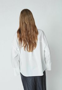 Chemise blanche oversize avec un ourlet incurvé et des manches longues, portée avec une jupe plissée noire. Tissu fluide et coupe décontractée.