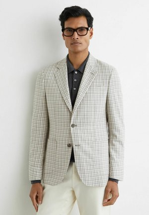 Mann mit schwarzer Brille, beigem Hahnentritt-Blazer über dunklem grauem Poloshirt und cremefarbenen Hosen, steht vor einer weißen Wand.