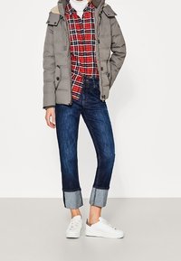 Grå polstret jakke med en hætte forsynet med pels, rød rutet skjorte, mørk denim jeans med oprullede hem, og hvide sneakers.