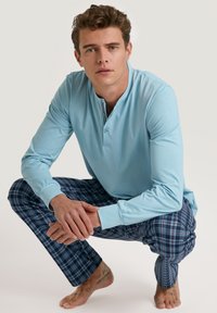 Lichtblauwe lange mouwen henley met twee knopen, gecombineerd met donkere geruite broek met blauwe, rode en witte vierkanten. Natuurlijke huidskleur.