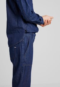 Personne portant une chemise et un pantalon en denim bleu foncé assortis, les mains jointes devant sur un fond clair uni.
