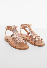 Pár plochých sandálů s řemínky v růžově zlaté metalické barvě, s přezkami na kotnících a hnědými podrážkami, vystavených na světlém pozadí.