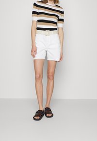 Person iført en beige, sort og hvid stribet kortærmet skjorte, hvide shorts med bælte og mørkebrune sandaler, der står foran en ensfarvet baggrund.