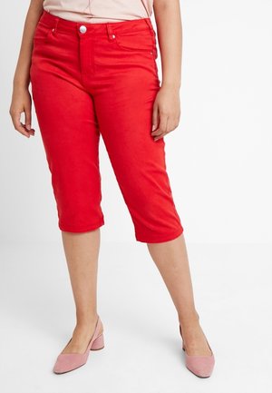 Femme portant un pantalon capri rouge vif et des chaussures roses à talons bas, debout devant un fond blanc uni.