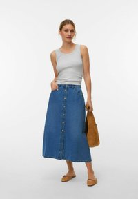 Sieviete valkā pelēku bezroku topu, pogām aiztaisāmu džinsa midi svārku, tur rokā brūnu zamšādas rokassomu un valkā bēšus dzīvokļus ar bantītēm.