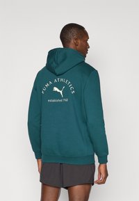 Sudadera color teal hecha de una tela suave, con un gran logo blanco de Puma y texto en la parte posterior. Incluye un capucho con cordón y puños acanalados.