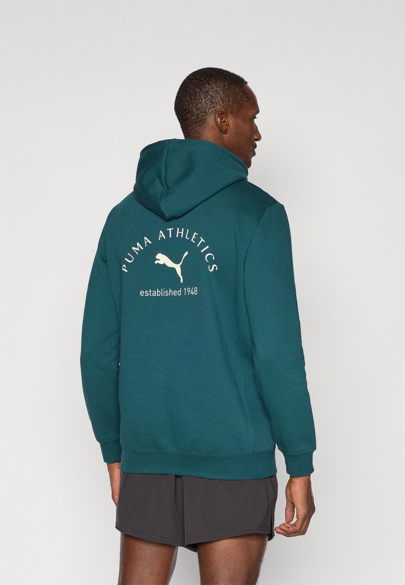 Sudadera color teal hecha de una tela suave, con un gran logo blanco de Puma y texto en la parte posterior. Incluye un capucho con cordón y puños acanalados.