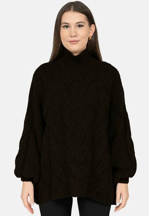 Woman with long brown hair wearing a black chunky knit sweater with a high neck and balloon sleeves, standing against a plain background.