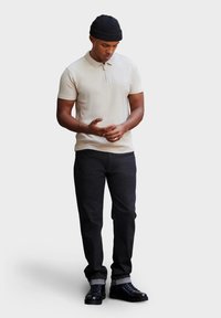 Beige polo-paita, jossa on vetoketju, yhdistettynä tummiin farkkuihin ja mustiin nauhakenkiin. Mallilla on päässään musta neulottu pipot.
