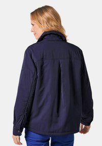 Dunkelblaue Jacke mit hohem Kragen, mit einer Falte hinten, elastischen Manschetten und Knopfdetails. Hergestellt aus glattem, leichtem Stoff.