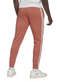 Red-orange joggers made of soft fabric feature three white stripes down the sides, tapered fit, and ribbed cuffs, paired with white sneakers.