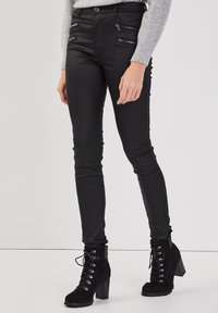 Jean skinny noir avec poches zippées à l'avant, coupe slim et texture lisse, associé à des bottines noires à lacets avec des talons épais.