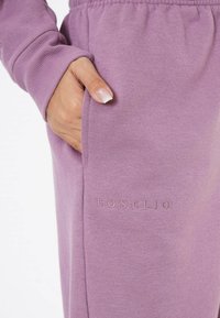 Lila Sweatpants mit weichem Material, ausgestattet mit einer Seitentasche und einem geprägten Logo "EOS ELIO" auf der Vorderseite.