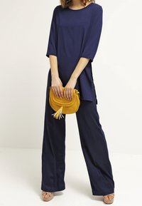 Mörkblå tunika med trekvartsärmar, avslappnad passform, sidoslitsar, som är ihop med vida byxor och en gul korsväska med fransar.