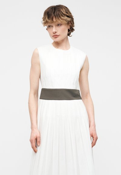 Νεαρή γυναίκα με κοντά κυματιστά μαλλιά, φοράει λευκό πλισέ φόρεμα χωρίς μανίκια και φαρδιά σκοτεινή ζώνη, στέκεται μπροστά σε απλό φωτεινό φόντο.