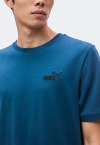 Άνδρας φοράει μπλε μπλούζα Puma με μικρό μαύρο λογότυπο Puma στο αριστερό στήθος, εμφανιζόμενος από τον λαιμό μέχρι το μέσο του κορμού.
