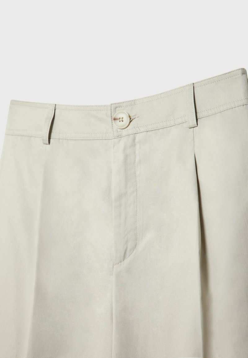 Pantaloni beige con davanti piatto, chiusura a singolo bottone, tasche laterali e dettagli plissettati. Realizzati in tessuto leggero con una texture liscia.