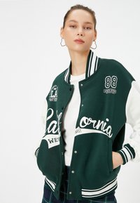 Zielona kurtka varsity z białymi rękawami, z napisem "California" i detalami w postaci naszywek. Żebrowany kołnierz i mankiety, zapięcie na napy.