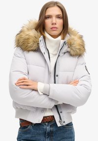 Doudoune grise clair avec capuche en fausse fourrure. Comprend une fermeture éclair, des poignets côtelés et un design matelassé. Portée par-dessus un col roulé blanc.